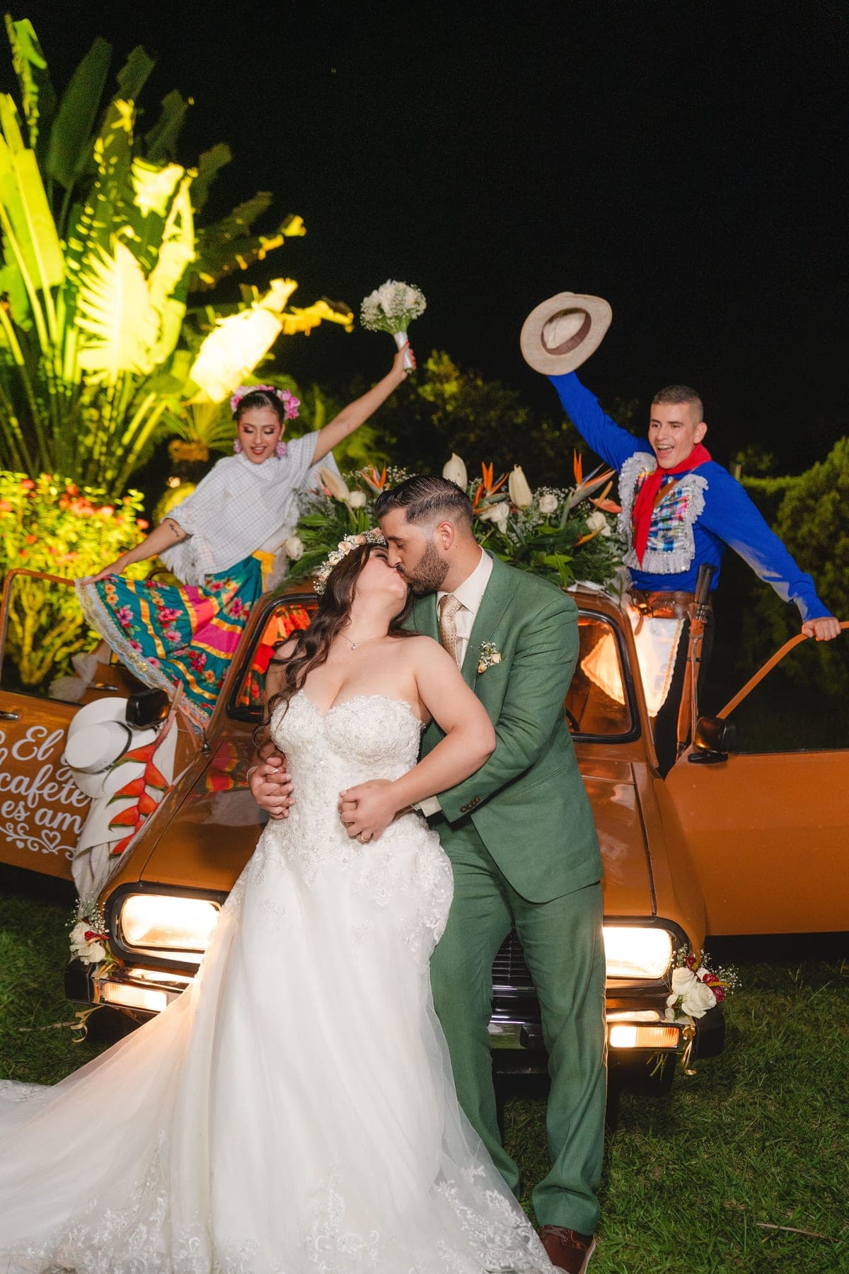 Novios se besan frente a un auto clásico mientras los invitados celebran